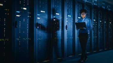 In Dark Data Center: Male IT Specialist Walks along the Row of Operational Server Racks, Uses Laptop for Maintenance. Concept for Cloud Computing, Artificial Intelligence, Supercomputer, Cybersecurity
