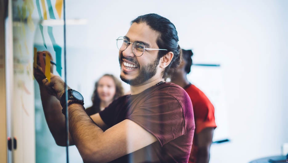 Side view of cheerful bearded man in glasses writing memos on sticky notes while creating plan on glass board in office with colleagues